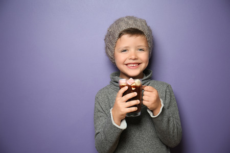 Cute little boy in warm clothes with glass cup of hot chocolate on color backgroundの写真素材