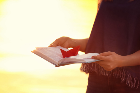 Woman holding open book with origami boat near river at sunsetの写真素材