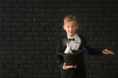 Cute little magician holding hat with rabbit against dark brick wallの写真素材