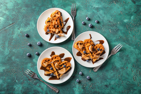 Heart shaped waffles with banana slices and chocolate sauce on color background