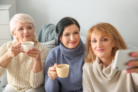 Beautiful mature woman drinking coffee while taking selfie at homeの写真素材