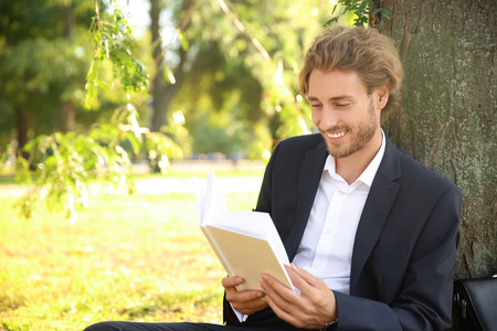 Handsome young businessman with book resting near tree in parkの写真素材