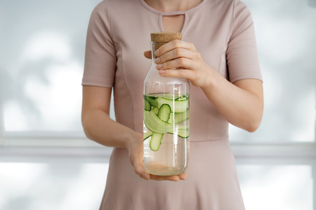 Woman holding bottle of fresh cucumber water on light background