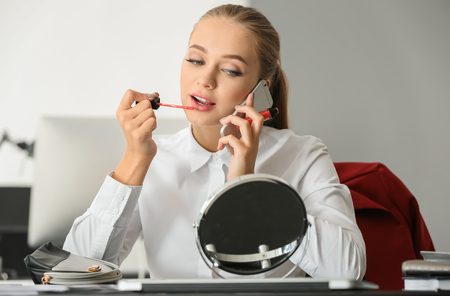 Beautiful female secretary applying makeup while talking on mobile phone in officeの写真素材