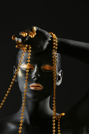 Beautiful woman with black and golden paint on her body holding beads against dark backgroundの写真素材