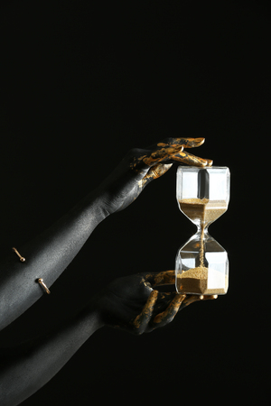 Woman with black and golden paint on her body holding hourglass against dark backgroundの写真素材