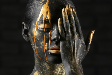 Beautiful woman with black and golden paint on her body against dark backgroundの写真素材