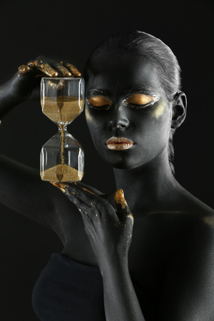 Beautiful woman with black and golden paint on her body holding hourglass against dark backgroundの写真素材