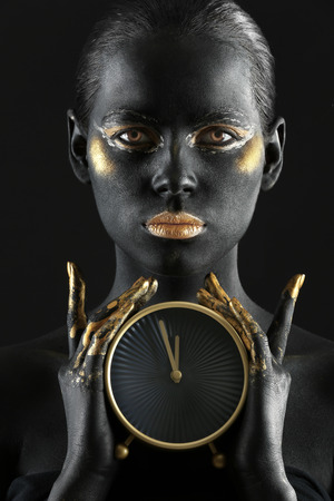 Beautiful woman with black and golden paint on her body holding clock against dark backgroundの写真素材