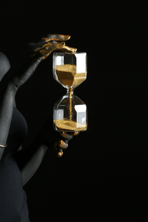 Woman with black and golden paint on her body holding hourglass against dark background
