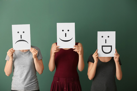 Young women hiding faces behind sheets of paper with drawn emoticons on color backgroundの写真素材