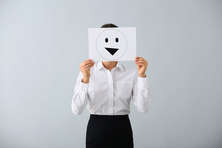 Young woman hiding face behind sheet of paper with drawn emoticon on light backgroundの写真素材