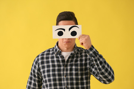 Emotional young man hiding face behind sheet of paper with drawn eyes on color backgroundの写真素材