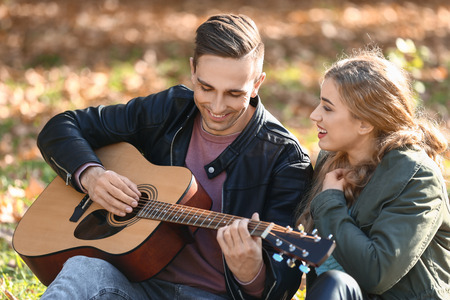 Handsome young man with guitar and his beloved resting in autumn parkの写真素材