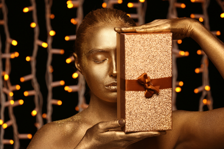 Beautiful young woman with golden paint on her body and gift box against defocused lightsの写真素材