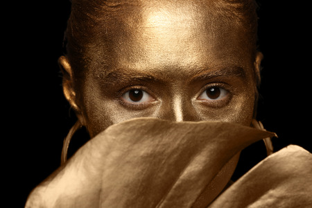 Beautiful young woman with golden paint on her body and tropical leaf against dark background,...