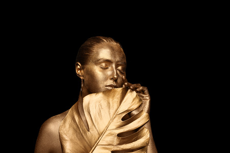 Beautiful young woman with golden paint on her body and tropical leaf against dark backgroundの写真素材