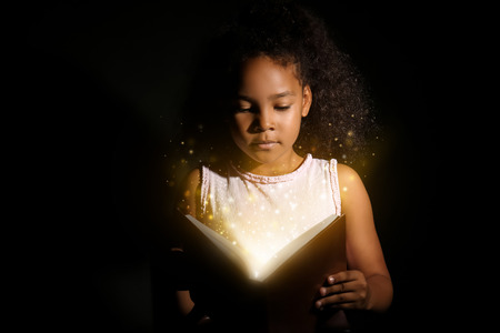 Little African-American girl reading magic book on dark backgroundの写真素材
