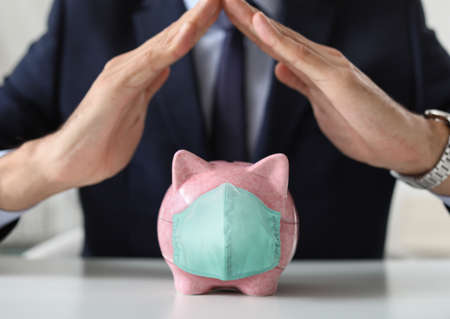 Male bank manager with piggy bank in medical protective mask sitting at table, closeup