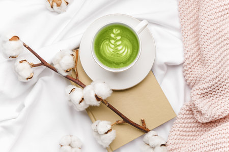 Composition with cup of matcha tea, beautiful cotton branch and book on bedの写真素材
