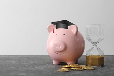 Piggy bank with graduation hat, coins and hourglass on light background. Concept of savings for educationの写真素材