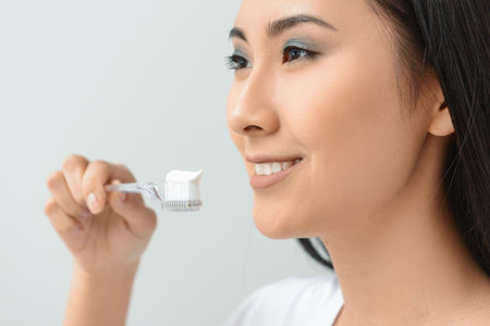 Beautiful Asian woman with toothbrush and paste on light background