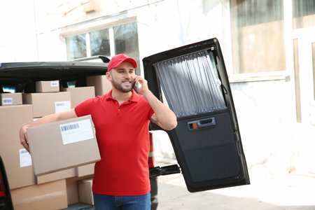 Delivery man talking by phone while standing near car with parcels