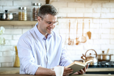 Handsome mature man reading book in kitchen at homeの写真素材