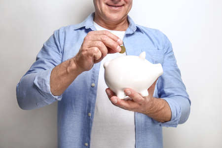 Handsome middle-aged man putting coin into piggy bank on light backgroundの写真素材