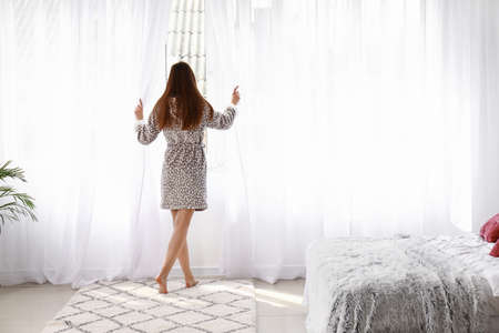 Young woman opening light curtains in bedroom at homeの写真素材