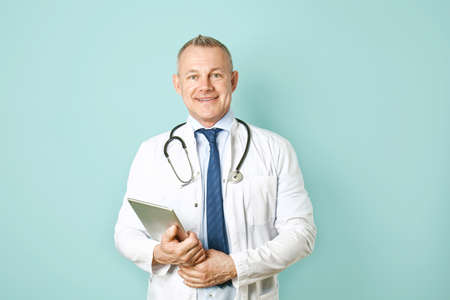 Handsome middle-aged doctor with tablet PC on color background