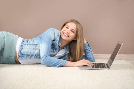 Beautiful young woman with laptop lying on floor near color wallの写真素材