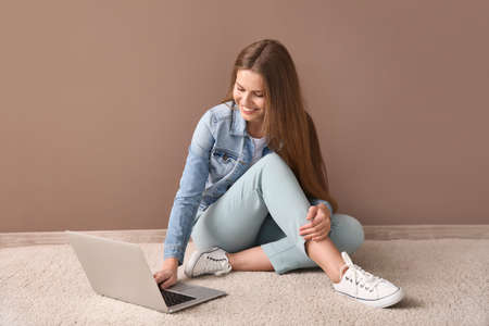 Beautiful young woman with laptop sitting on floor near color wallの写真素材