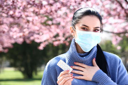 Young woman with antihistamine pills wearing protective mask near blooming tree