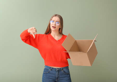 Young woman with cardboard box showing thumb-down on color background