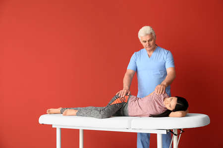 Mature physiotherapist working with young Asian woman on color background