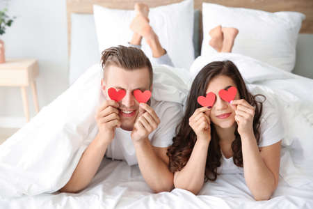 Happy young couple with paper hearts lying in bed