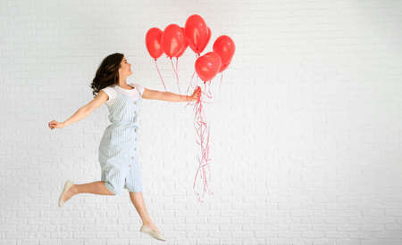 Jumping young woman with heart shaped air balloons near white brick wallの写真素材
