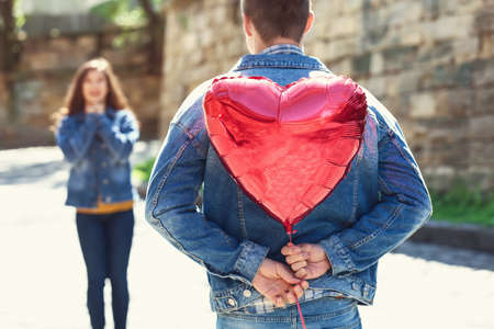 Young man hiding heart shaped air balloon for girlfriend behind his back outdoorsの写真素材