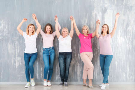 Happy women of different ages with pink ribbons near gray wall. Breast cancer concept