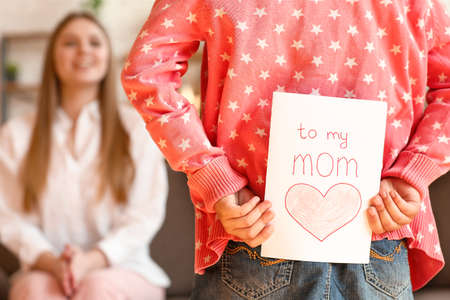 Little daughter hiding greeting card for her mother behind back at home
