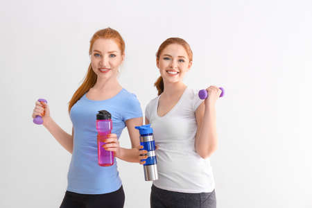Beautiful sporty young women with bottles of water and dumbbells on light background