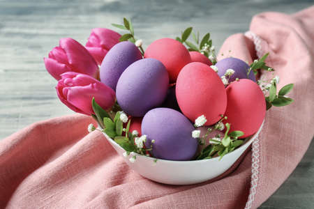 Bowl with beautiful Easter eggs and flowers on gray table