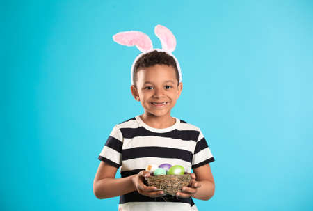 Little African-American boy with Easter eggs and bunny ears on color background