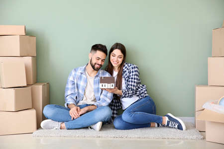 Young couple with belongings and model of house sitting near color wall