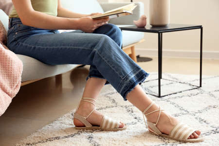 Young woman in stylish jeans pants reading book on sofa at homeの写真素材