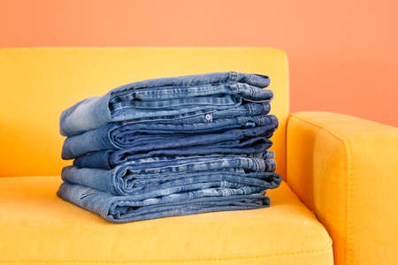 Stack of jeans pants in armchair on color background