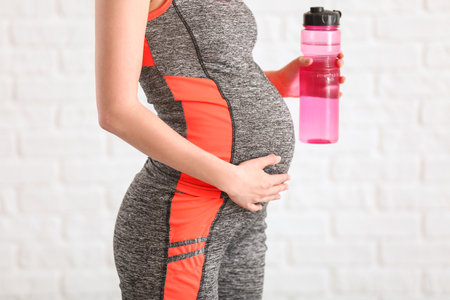 Beautiful sporty pregnant woman drinking water near white brick wallの写真素材