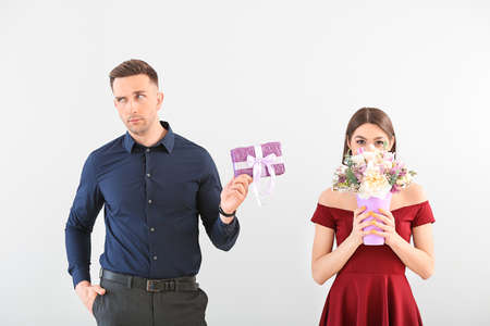 Displeased young man with gift box and happy woman with bouquet of beautiful flowers on light backgroundの写真素材