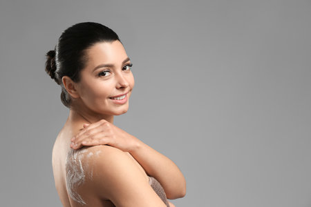Beautiful young woman applying body scrub against gray background